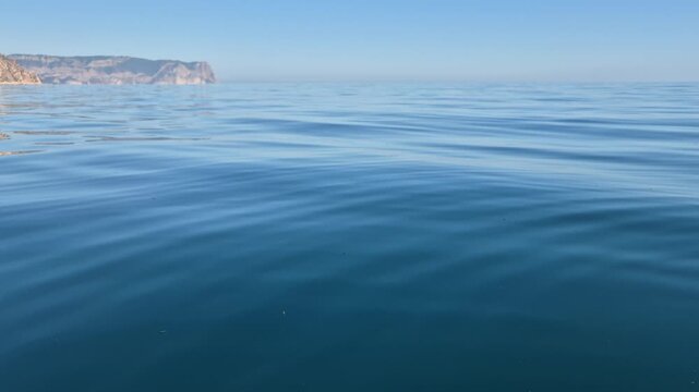 Calm sea water, peaceful blue ocean waves ripple with distant majestic cliffs under a clear horizon sky
