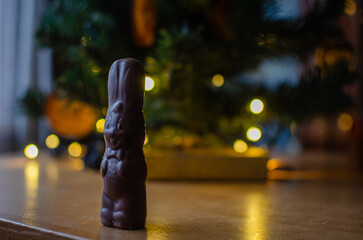 A charming dark chocolate bunny stands tall on a wooden table, dressed with a little bow tie and a cute face. It poses in front of a beautifully lit Christmas tree with warm bokeh lights, creating a c