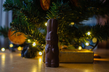 A charming dark chocolate bunny stands tall on a wooden table, dressed with a little bow tie and a cute face. It poses in front of a beautifully lit Christmas tree with warm bokeh lights, creating a c