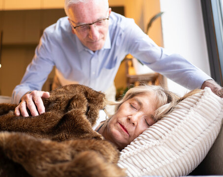 Senior husband covering his wife with a blanket, showing care and love during her nap