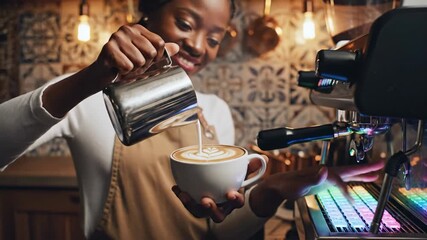 Skilled African American female barista smiling while pouring steamed milk to create beautiful latte art in a modern, warmly lit coffee shop