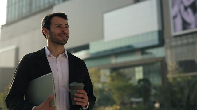 Portrait of happy Hispanic businessman with coffee cup to go and laptop walks across modern city slow motion. Young office employee in formal suit expresses confidence in downtown
