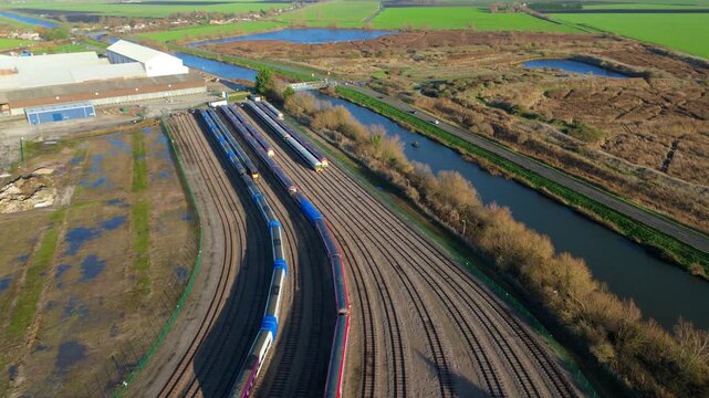 Aerial view of trains on tracks, contrasted by the river and green fields beyond, creating a striking juxtaposition of industry and nature, Ely, England, United Kingdom.