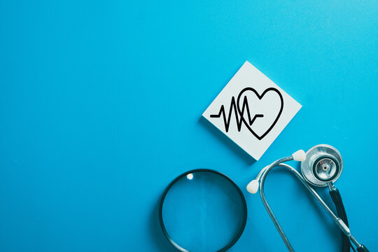 Medical stethoscope and magnifying glass near heart pulse icon on note on blue background. Concept of healthcare checkup, cardiology diagnosis, life insurance policy, and wellness protection. - Powered by Adobe