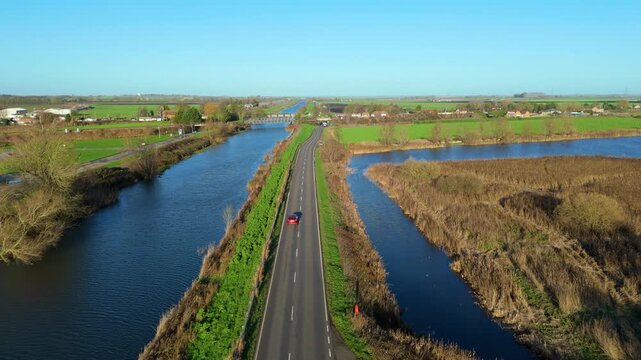 Aerial view of a solitary car driving along a road bisecting waterways, with green fields in the distance, creating a vibrant contrast, Ely, England, United Kingdom.