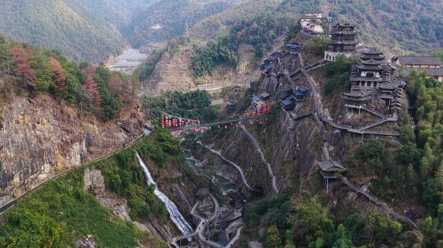 Aerial view of Wangxian Valley's traditional buildings perched on rocky cliffsides, connected by winding pathways and a vibrant red bridge, Wangxian Valley, China.
