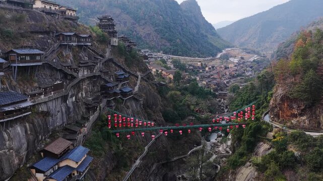 Aerial view of Wangxian Valley showcasing architecture clinging to the rugged cliffs and a bridge adorned with vibrant red lanterns, Wangxian Valley, China.
