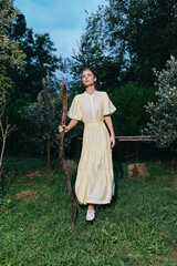 Fototapeta premium Woman in a pale yellow dress stands in a grassy garden holding a wooden staff, surrounded by trees under soft evening light, conveying calmness and rural charm.