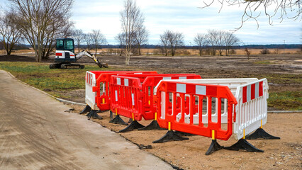 Construction site with yellow mini excavator and orange safety barriers on dirt field near road, autumn birch trees under cloudy sky, roadwork equipment and traffic control scene.
