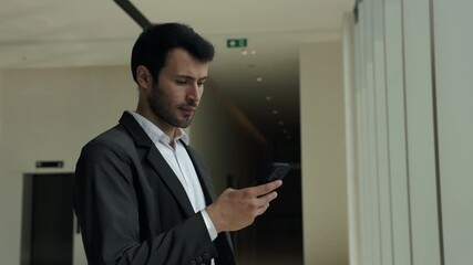 Portrait of bearded Hispanic man corporate employee looks at smartphone in office building. Latin manager in formal suit reads message on modern mobile phone in business center