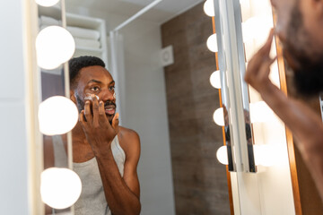 Black man applying face cream while looking in a bathroom mirror with lights, focused and confident...