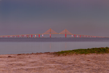 The Sunshine Skyway Bridge rises gracefully against the night sky, illuminated in a striking display of color and light. Reflections shimmer across the calm water below, enhancing the bridge&rsquo;s elegant