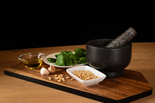 Still life of a setup with  ingredients and kitchen tool to make pesto, such as basil, garlic, olive oil and pine nuts. Black mortar as tool to grind the ingredients