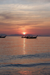 Obraz premium Two boats approach the shore at twilight, floating on soft pink waters with the sun descending behind distant mountains. 