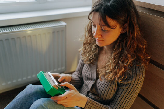 Young woman sitting comfortably on the floor, playing the kalimba, a small musical instrument, next to the radiator, enjoying a warm and cozy atmosphere at home during winter