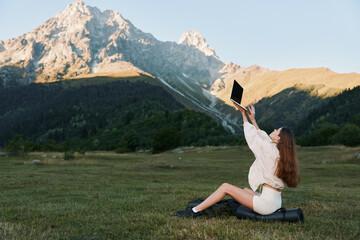 A joyful woman sits on a grassy meadow beneath rugged mountain peaks, lifting a laptop to capture a playful selfie against a clear blue sky.