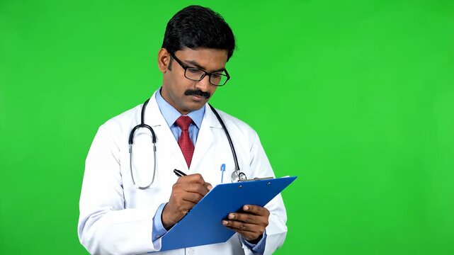 Professional Indian Male Doctor Taking Notes on Chart Against Green Screen