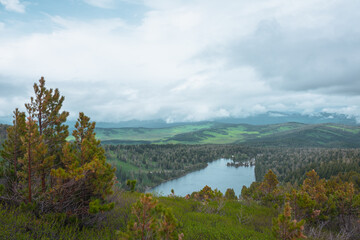 Scenic top view from green lush thickets and coniferous trees on mountain pass to big wood lake against hilly vastness far away in low clouds. Long forest lake and sunlit woody hills under cloudy sky.