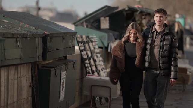 A young couple walks along the Seine River in Paris France The clip shows a medium shot of the couple walking past old book stalls and buildings The camera is handheld giving a natural feel to the sce