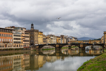 Colorful riverside buildings and Ponte Vecchio bridge reflecting in the Arno River under a dramatic cloudy sky in Florence, Italy, with a flying seagull and scenic hills in the background