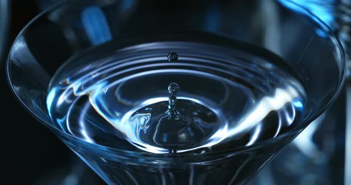 Slow motion 1000 fps shot of a drop falling into a clear cocktail in a martini glass. Blurred elegant bar background, soft intimate lighting, luxury nightlife and premium drink concept.