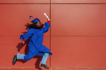 Female student jumping with diploma wearing graduation gown and cap against red wall