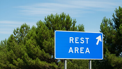 Blue rest area sign with white arrow pointing right against green trees and blue sky, indicating a place to stop and relax during travel.