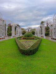Symmetrical garden pathway framed by white decorative arches leads to heart-shaped hedge with small plant in center, surrounded by green lawns under cloudy sky. Sochi, Russia - December 13, 2025