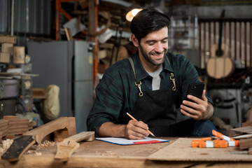 Carpenter using smartphone and taking notes in woodworking workshop, small business craftsman