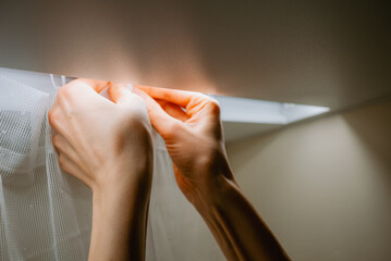 Woman's hands meticulously hanging new white tulle curtains onto a ceiling mounted track, focusing...