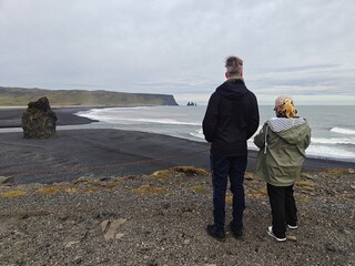 Couple looking at Reynisfjara black sand beach in Iceland. View of volcanic coast with basalt sea stacks and Atlantic ocean under moody sky. Scenic Nordic nature and travel concept.