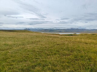 Obraz premium Panoramic Icelandic landscape with a vast golden grass field, a calm lake, and distant snowy mountains under a cloudy sky.