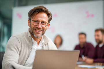 Smiling mature businessman with glasses sitting at a laptop in a modern office with colleagues collaborating in the background during a meeting session