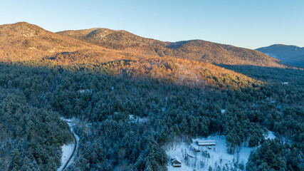 An aerial winter landscape of the Adirondack Mountains at sunrise. Golden light hits the snowy peaks above a frost-covered evergreen forest.