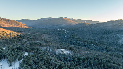 An aerial winter landscape of the Adirondack Mountains at sunrise. Golden light hits the snowy peaks above a frost-covered evergreen forest.
