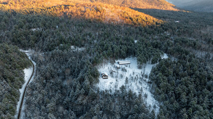 An aerial winter landscape of the Adirondack Mountains at sunrise. Golden light hits the snowy peaks above a frost-covered evergreen forest.
