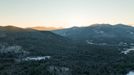 An aerial winter landscape of the Adirondack Mountains at sunrise. Golden light hits the snowy peaks above a frost-covered evergreen forest.