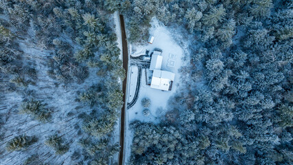 A top-down aerial view of a modern white house nestled in a snow-covered evergreen forest. A single dark road cuts vertically through the dense, frosty trees.