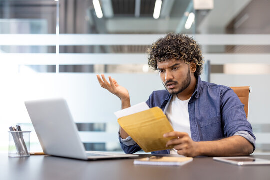 A worried young Indian man sits at his desk in the office and looks disappointedly at the letter he received