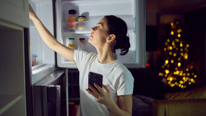 Woman choosing food from open refrigerator while holding smartphone at home. Concept of lifestyle ads, food delivery promotion, household services marketing and modern home consumption visuals.