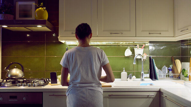 Rear view of woman cooking at kitchen counter with smartphone support. Concept of lifestyle ads for digital services, mobile apps, online platforms and modern home communication in promo campaigns. - Powered by Adobe