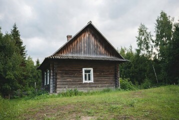 A traditional old wooden log house on the edge of a forest on a summer day. A rural dacha surrounded by green trees and grass under an overcast sky.
