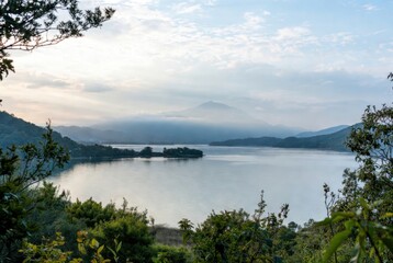 High-angle view of a calm lake with a small island, surrounded by lush green hills and a misty mountain peak in the distance. Serene natural landscape under a soft overcast sky at dawn.