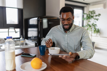 African American man sitting at a kitchen table, having breakfast and drinking coffee while using a digital tablet at home, relaxed yet focused, modern remote work and everyday lifestyle concept