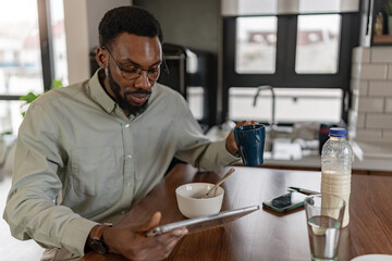 African American man sitting at a kitchen table, having breakfast and drinking coffee while using a digital tablet at home, relaxed yet focused, modern remote work and everyday lifestyle concept