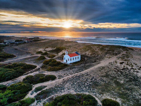 Aerial view of a charming white church with a red roof stands amidst windswept dunes as the sun bursts through dramatic clouds, painting the scene with golden light, Labenne, France.
