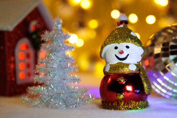 A snowman between a ball and a small Christmas tree with a house on a blurred background with yellow lights