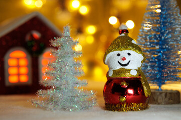 A snowman between a white and blue Christmas tree with a small red house on a blurred background with yellow lights