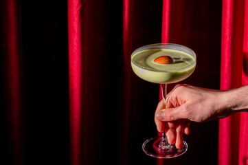Hand holds a vibrant green matcha cocktail in a coupe glass against a dramatic red velvet curtain background, creating a moody, luxurious bar atmosphere