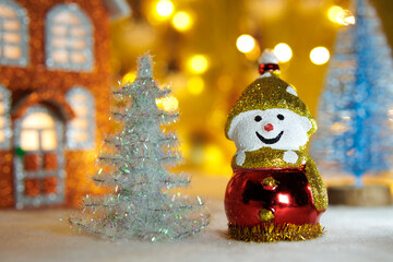 A snowman between a white and blue Christmas tree with an orange house on a blurred background with yellow lights